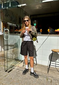 Jaqueta com estampado de leopardo, t-shirt branca, calções pretos plissados e sapatos de plataforma pretos com meias brancas; de pé à porta de vidro.