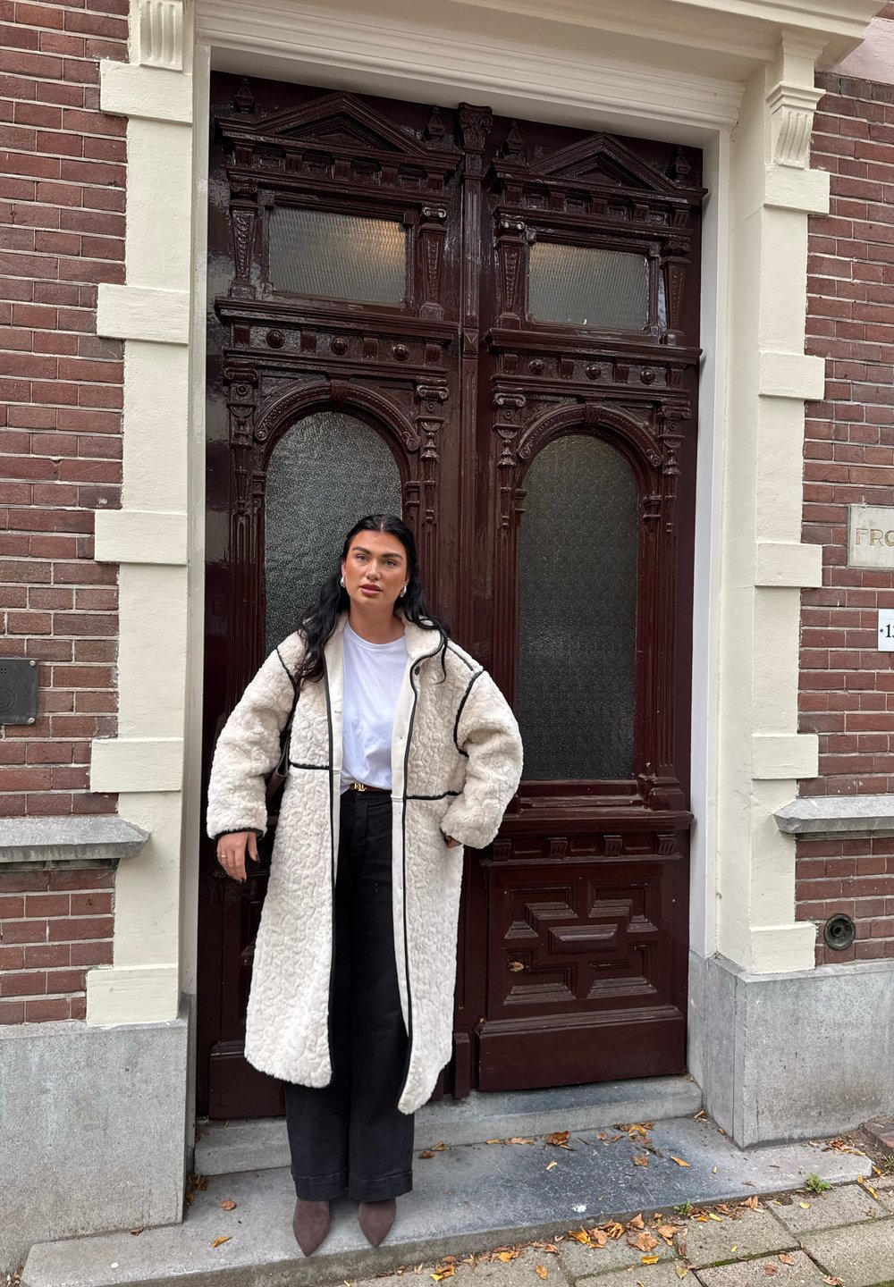 Casaco de pelagem oversized em creme com acabamentos pretos, usado por cima de uma camisa branca e calças pretas. Ocupa uma porta com detalhes em madeira ornamentados.
