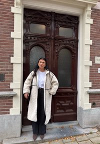 Casaco de pelagem oversized em creme com acabamentos pretos, usado por cima de uma camisa branca e calças pretas. Ocupa uma porta com detalhes em madeira ornamentados.