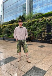 Täiskasvanud mees, kes kannab ruudulist pruuni särki, kamuflaažiga lühikesi pükse ja beeže jooksujalatseid, millel on värvilised aktsendid, seismas kivist sillutisel.