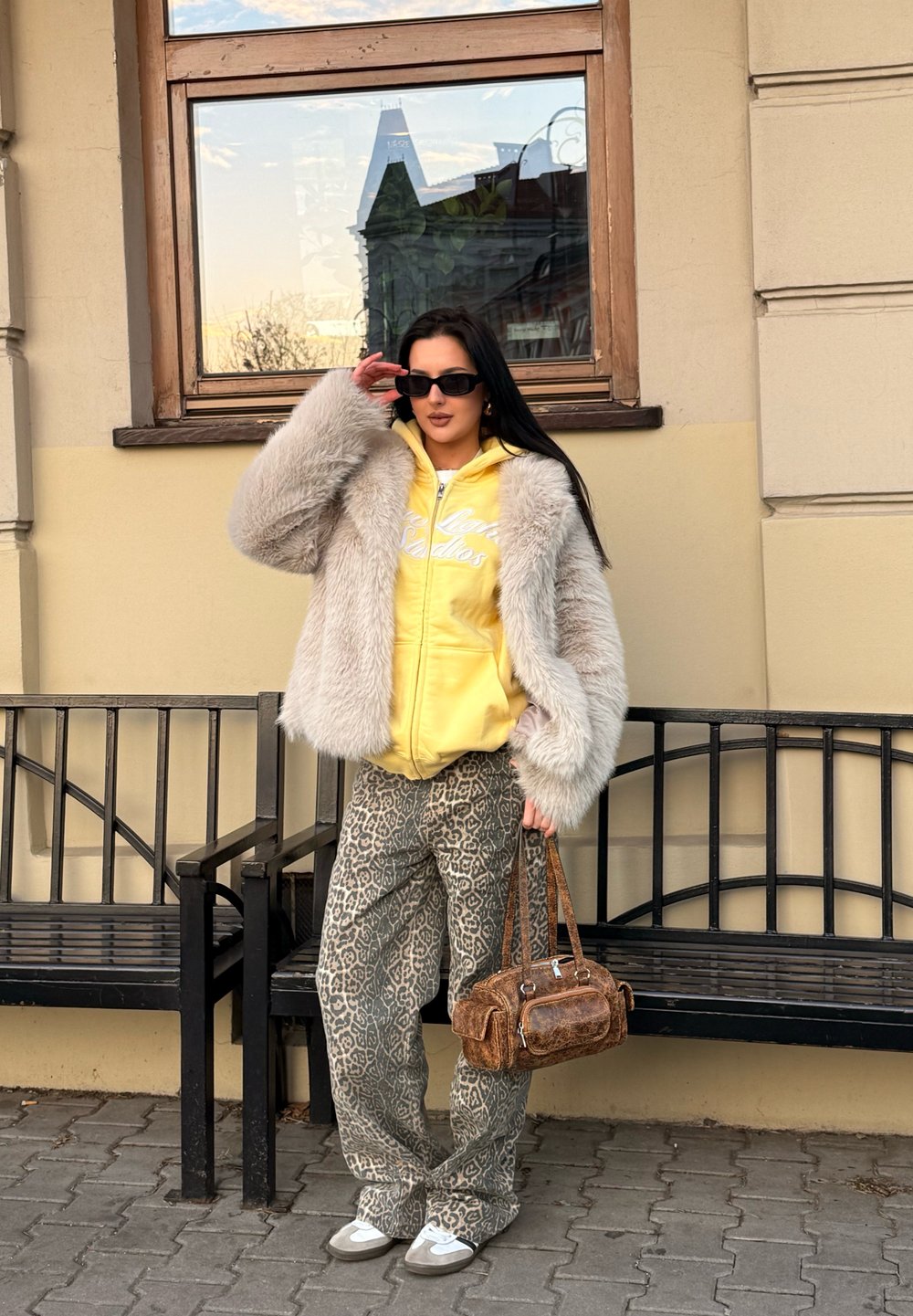 Casaco de peluche cinza claro, hoodie amarelo com letras brancas, calças de padrão de leopardos e uma bolsa castanha com detalhes. Óculos de sol pretos.