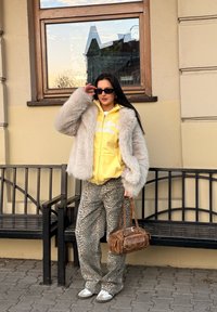 Casaco de peluche cinza claro, hoodie amarelo com letras brancas, calças de padrão de leopardos e uma bolsa castanha com detalhes. Óculos de sol pretos.