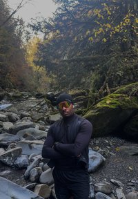 Zwarte mouwloze bodywarmer over een nauwsluitend shirt, zwarte muts en reflecterende zonnebril. Op de achtergrond zijn rotsen, groenblijvende planten en een rivier te zien.