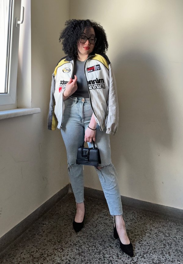 Giacca bomber grigia e gialla con loghi, camicia scura aderente, jeans chiari strappati, tacchi alti neri e una piccola borsa nera.