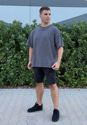 Camiseta de algodón gris de gran tamaño con cuello redondo, combinada con pantalones cortos de mezclilla negros desgastados y zapatillas negras, de pie sobre una superficie pavimentada.