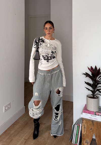 Camiseta blanca de manga larga con detalle gráfico negro, combinada con pantalones de chándal grises desgastados. Botas negras de punta completan el conjunto. Accesorios mínimos visibles.
