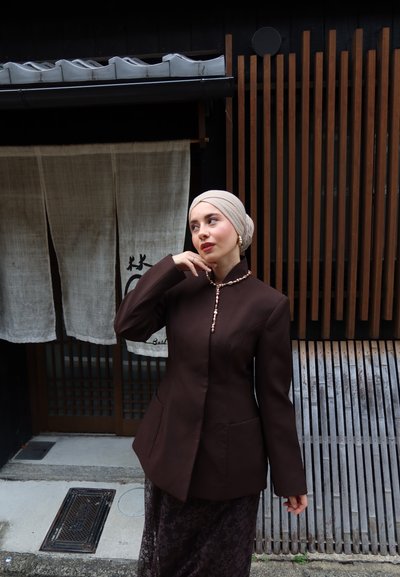 Chaqueta marrón a medida con cuello alto, cierre de botones y bolsillos frontales. Llevada sobre una falda oscura con estampado, contra un fondo de madera y tela.