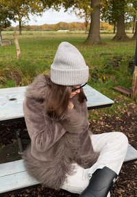 Rudas flisinis švarkas su tekstūruota priekyje, pilkos spalvos megztinė kepurė, pilkos sportinės kelnės ir juodi guminiai batai, sėdintys prie lauko stalo žolėtoje vietoje.