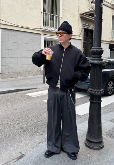Chaqueta bomber negra con cremallera frontal, pantalones de pierna ancha grises, gorro negro, gafas de sol de gran tamaño y sosteniendo una bebida helada clara.