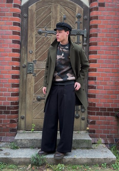 Abrigo verde oliva de doble botonadura, suéter con patrón de camuflaje, pantalones anchos negros y zapatos marrones, posando frente a una puerta de madera y una pared de ladrillos.