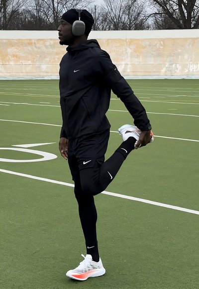 Ropa deportiva de Nike en negro, que incluye una chaqueta con cremallera y pantalones ajustados. La persona se estira en un campo verde, usando zapatillas blancas y naranjas.