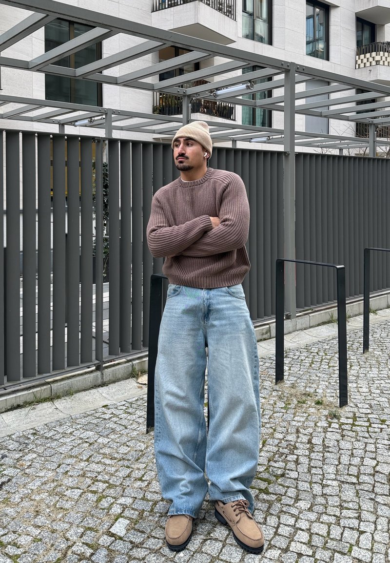 Homme portant un bonnet beige, un pull marron, un jean large bleu clair et des chaussures marron clair, debout bras croisés sur un chemin pavé devant un bâtiment moderne.
