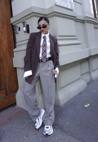 Kobieta ubrana w za dużą brązową marynarkę w prążki, białą koszulę z wydłużonymi mankietami, wzorzysty krawat, szare szerokie spodnie, białe tenisówki i czarne okulary przeciwsłoneczne.