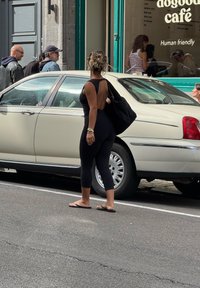 Žena v čiernom oblečení a sandáloch stojí pri bielom aute na ulici; okno kaviarne so zobrazenými ľuďmi v pozadí.