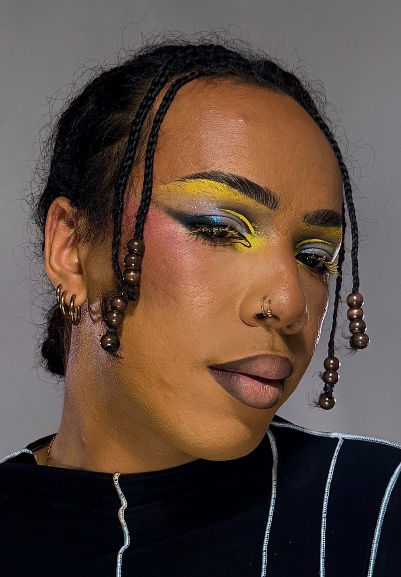 Person with braided hair adorned with beads, bold yellow and blue eye makeup, nose ring, multiple earrings, and black striped top.