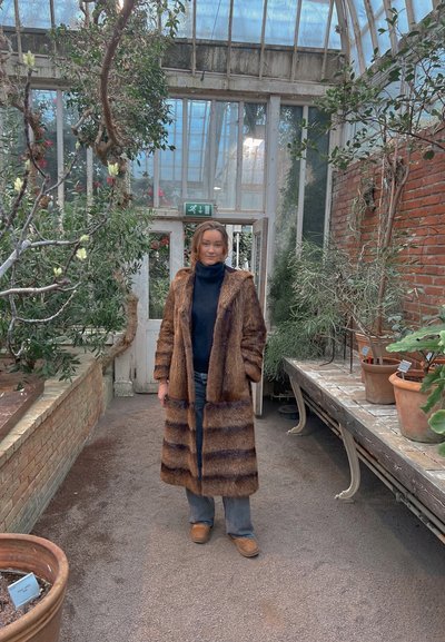 Abrigo de pelaje sintético marrón con patrón a rayas, combinado con un cuello de tortuga negro y jeans azules. Escenario de invernadero interior con plantas y paredes de ladrillo.