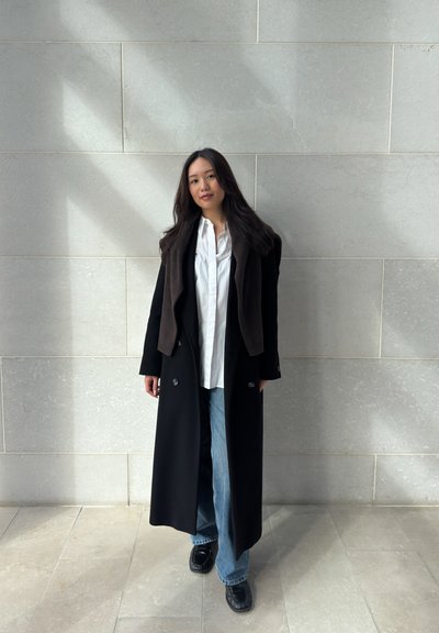 Mujer con cabello largo y oscuro, que lleva un abrigo negro largo, camisa blanca, jeans azules y zapatos negros, de pie contra una pared de piedra clara.