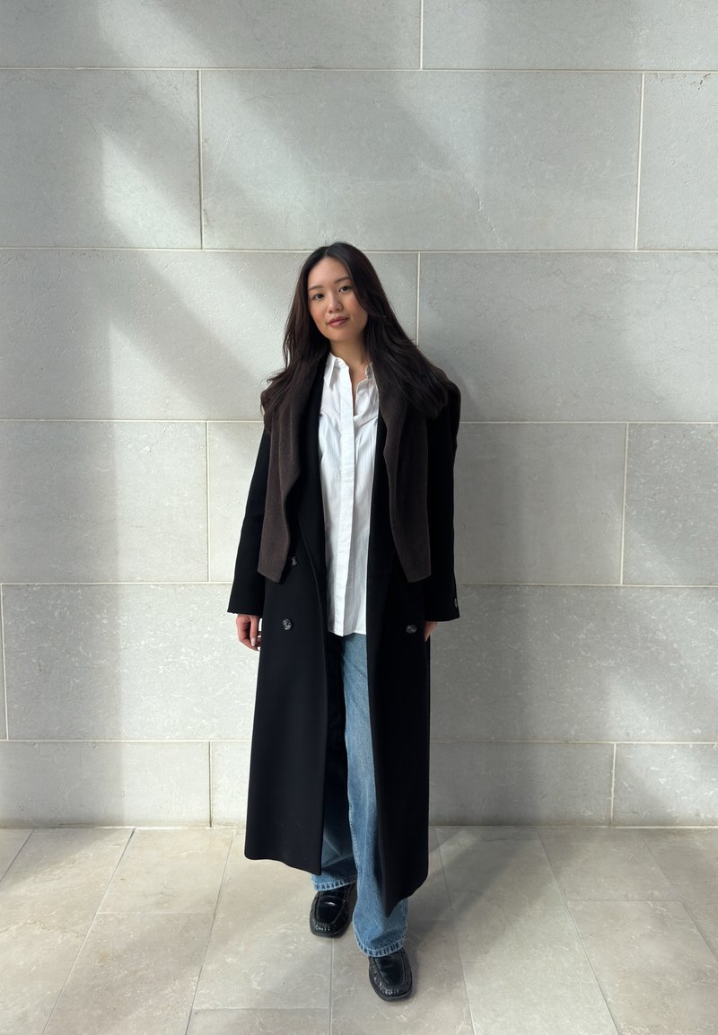 Femme aux longs cheveux foncés portant un long manteau noir, une chemise blanche, un jean bleu et des chaussures noires, debout contre un mur de pierre clair.
