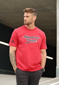 Camiseta de algodón roja con el logo de "Manchester United" en script negro, mangas cortas y un logo negro de Adidas en la manga izquierda.