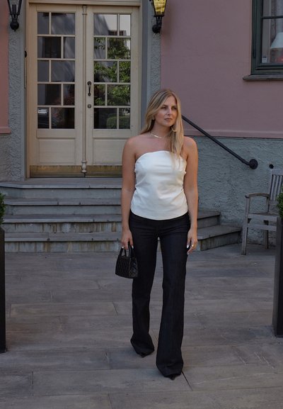 Top blanco sin tirantes, pantalones negros de campana, llevando un pequeño bolso negro acolchado. Textura suave, silueta ajustada, entorno al aire libre con escaleras.