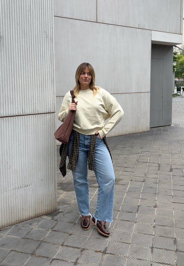 Cream sweatshirt, blue wide-leg jeans, brown plaid shirt tied around the waist, brown sling bag, and dark shoes. Textured grey wall in the background.