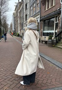 Long cream coat with side pockets, grey scarf, loose dark patterned trousers, and white slip-on shoes, standing on a cobbled street.