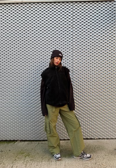 Mujer con gorro negro y chaqueta, pantalones cargo oliva y zapatillas, de pie frente a una pared metálica plateada texturizada al aire libre.