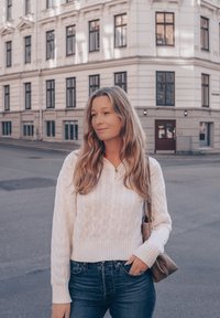 Valkoinen palmikkoneulepaita pitkillä hihoilla ja V-kaula-aukolla, yhdistetty tummansinihin farkkuihin. Ruskea olkalaukku. Kaupunkitausta.