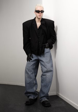 Persona con cabello corto y rubio, vistiendo un blazer negro oversize, guantes negros, gafas de sol ovaladas, vaqueros anchos azules y zapatos negros, de pie en el interior.