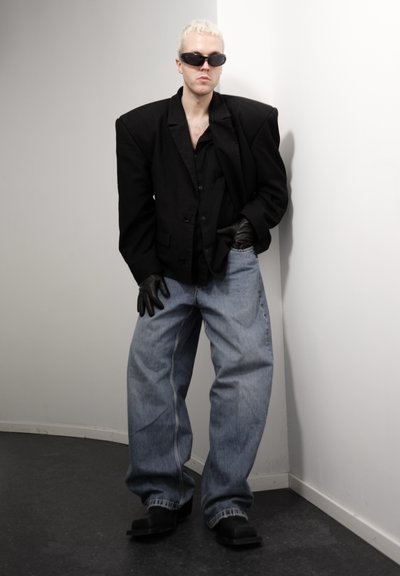 Persona con cabello corto y rubio, vistiendo un blazer negro oversize, guantes negros, gafas de sol ovaladas, vaqueros anchos azules y zapatos negros, de pie en el interior.