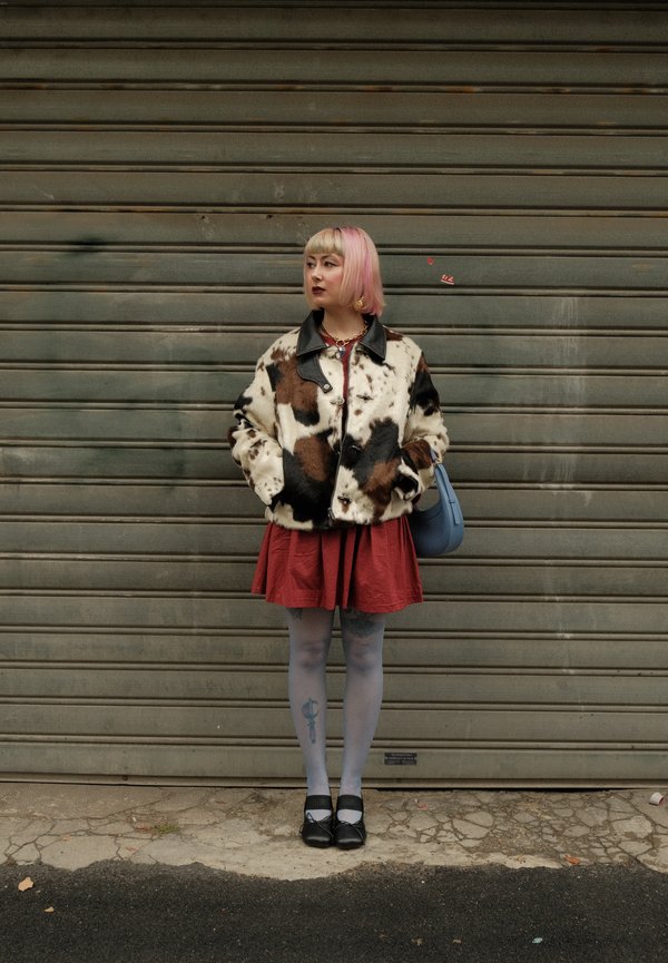 Una persona con el pelo rosa y rubio está de pie delante de una persiana metálica. Lleva una chaqueta estampada, una falda roja y medias, y sostiene un bolso azul.