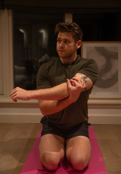 Una persona se arrodilla sobre una esterilla de yoga rosa, vistiendo una camiseta verde oscuro y pantalones cortos, estirando su brazo contra su codo en una habitación bien iluminada.