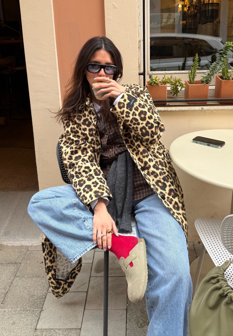 Personne portant un manteau à imprimé léopard, un jean bleu, des chaussettes rouges et des chaussures beiges, assise à l'extérieur d'un café en buvant dans une petite tasse.