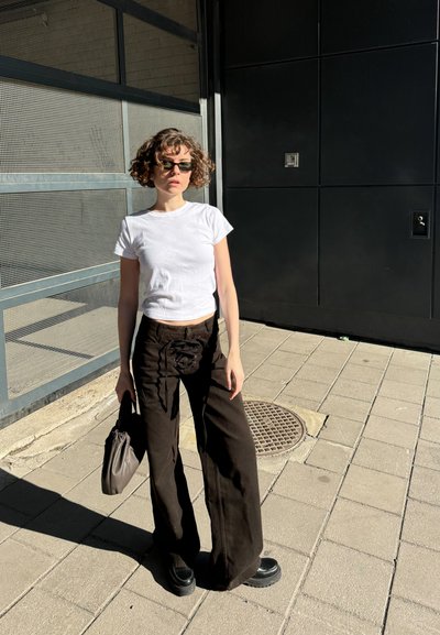 Camiseta corta blanca, pantalones de pierna ancha marrón oscuro con detalle de encaje, zapatos negros de suela gruesa y un pequeño bolso gris. La luz solar brillante proyecta sombras.