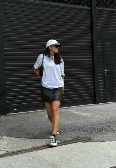 Camiseta blanca de gran tamaño, falda de cuero negra con cremallera, zapatillas negras con suela blanca y una gorra bicolor. De pie en una acera.