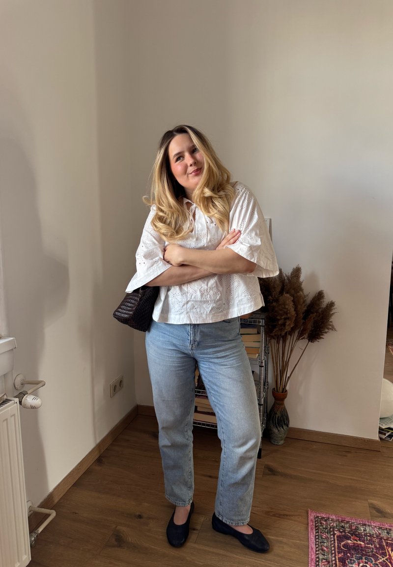 Vrouw met lang blond haar staat met gekruiste armen, draagt een witte blouse, lichtblauwe spijkerbroek en zwarte schoenen in een kamer met houten vloer en planten.