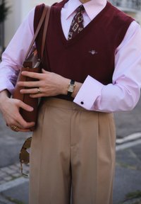 Bordo džemper bez rukava preko svijetloružaste košulje, bež plisirane hlače, smešnata kožna torba, zlatni sat i vidljiva kravata s uzorkom.