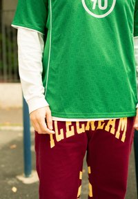 Maillot de sport vert avec un motif à pois et des finitions blanches, associé à un pantalon de survêtement bordeaux arbore des lettres jaunes. Geste de la main visible.