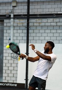 Mees valge spordisärgi ja mustade lühikeste pükstega lööb rohelise käepidemega reketiga kollase palli spordirajatises, millel on tellisseinad.