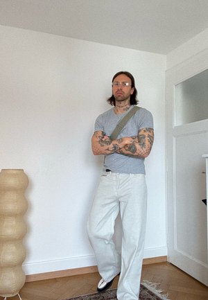 Hombre con tatuajes, gafas, camiseta gris, pantalones blancos y bolso de hombro, de pie con los brazos cruzados en una habitación minimalista.