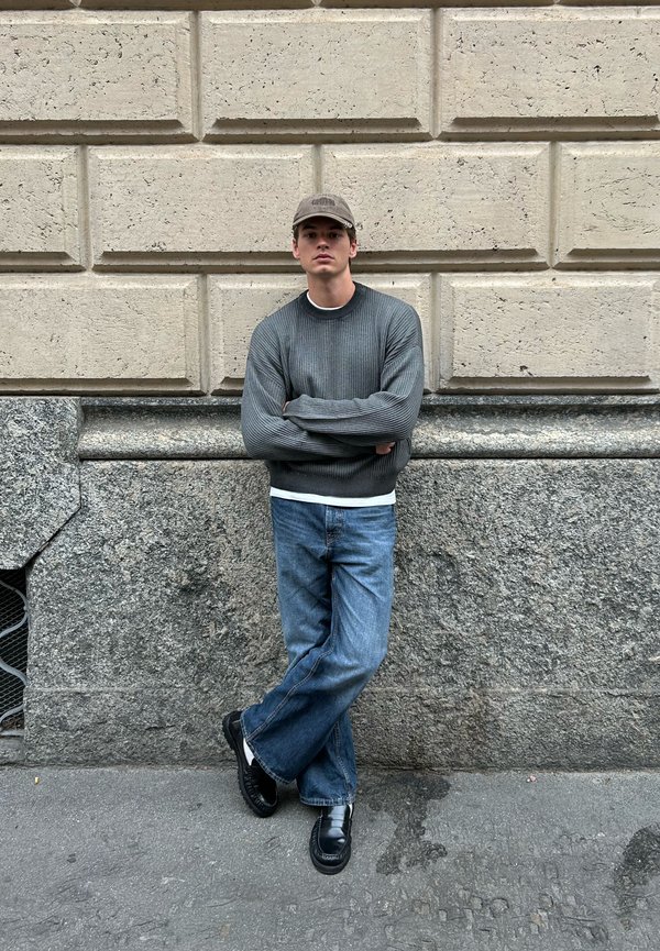 Junger Mann in grauem Pullover, blauen Jeans, schwarzen Schuhen und beigem Hut steht mit verschränkten Armen vor einer strukturierten Steinmauer.
