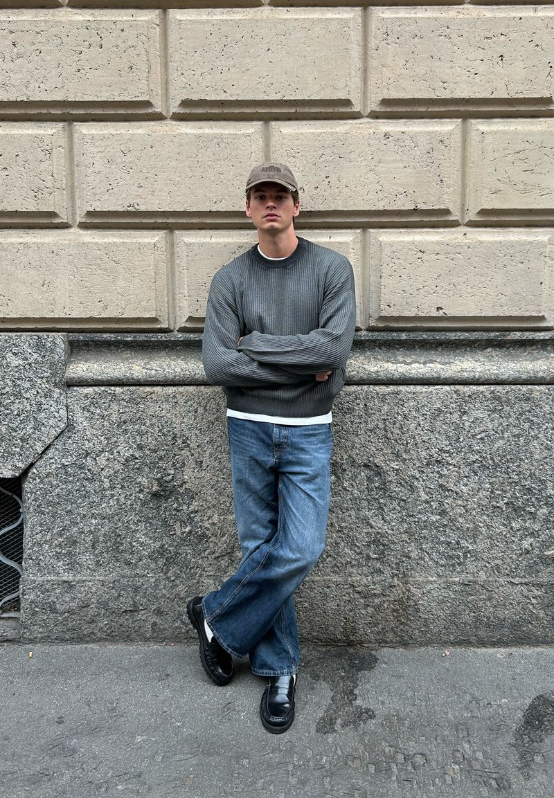 Giovane uomo che indossa un maglione grigio, jeans blu, scarpe nere e un berretto beige, si trova con le braccia incrociate contro un muro di pietra testurizzato.