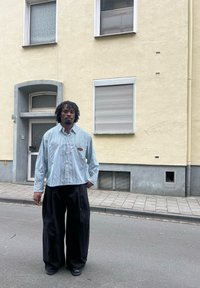 Camicia a righe blu e bianche con bottoni abbinata a pantaloni neri a gamba larga, posizionata davanti a una parete gialla e a una porta grigia.