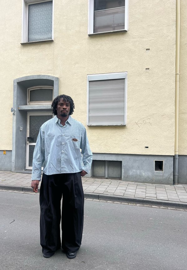 Gestreifte blau-weiße Button-Up-Bluse, kombiniert mit schwarzen weiten Hosen, vor einer gelben Wand und einer grauen Tür.