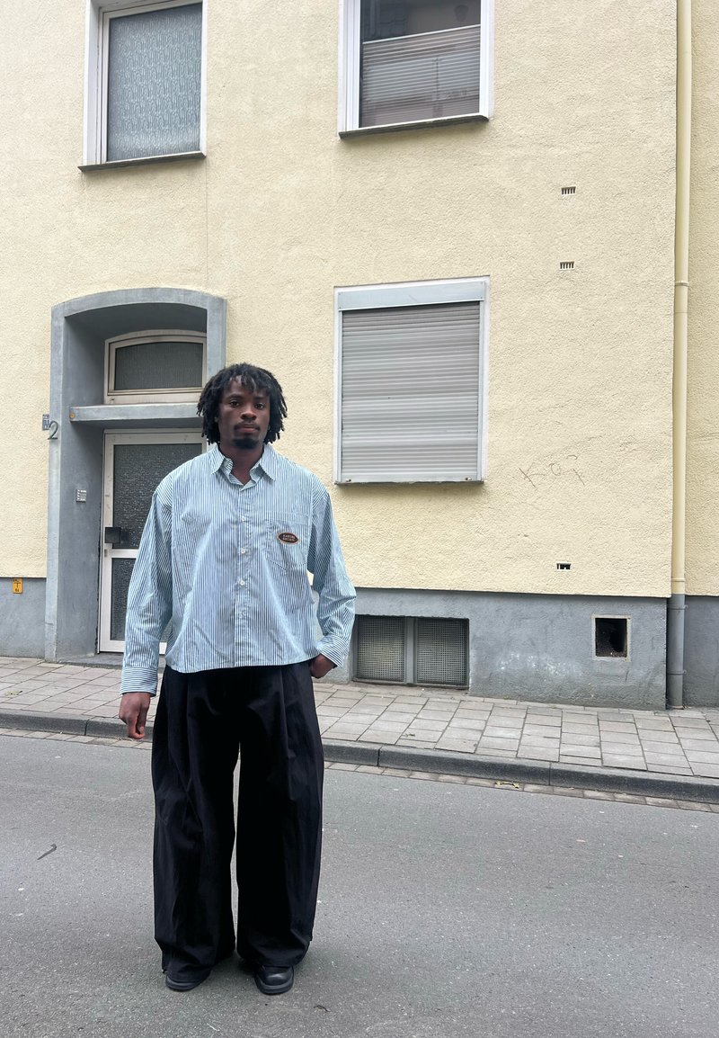 Gestreept blauw-wit button-up shirt gecombineerd met zwarte wijde broek, in een setting tegen een gele muur en een grijze deur.