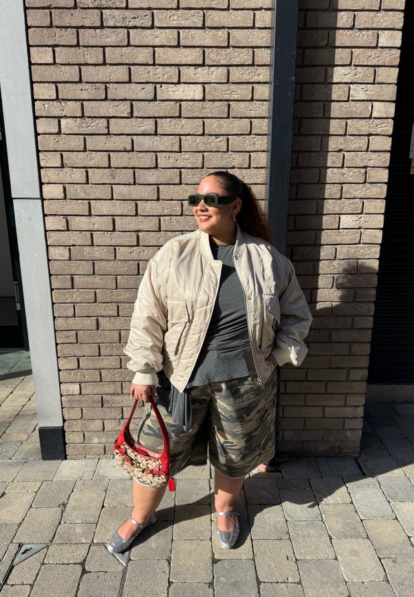 Una mujer con gafas de sol, una chaqueta color crema, pantalones cortos de camuflaje y sandalias sostiene un bolso rojo delante de un edificio de ladrillo.