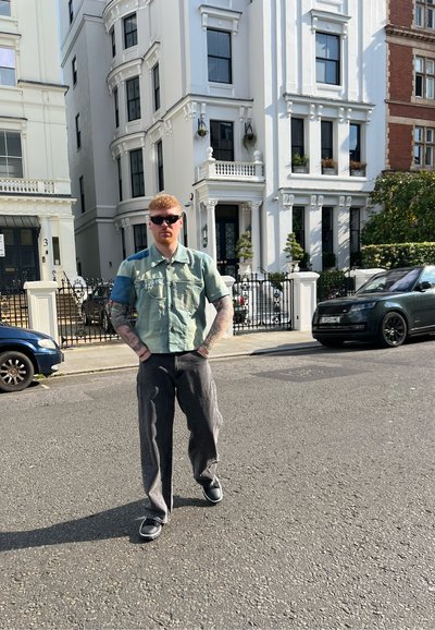 Camisa casual verde menta de manga corta, pantalones grises holgados y zapatillas negras. Lleva puestas gafas de sol, de pie cerca de edificios urbanos y coches.