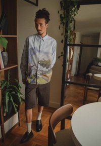 Overhemd met knopen en verticale strepen, met een kleurrijke landschapafdruk op de voorkant, gecombineerd met een donkere short en zwarte schoenen. Binnenomgeving.