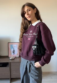 Vino rdeč pulover z belim ovratnikom z logotipom in britansko zastavo, združen s sivimi hlačami. Črna torba s trakcem in srebrnimi poudarki.