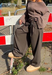 Maglione lavorato a maglia marrone over size con tasca, abbinato a pantaloni larghi scuri. Sneakers beige con platform completano l'outfit. Lo sfondo presenta barriere di sicurezza.